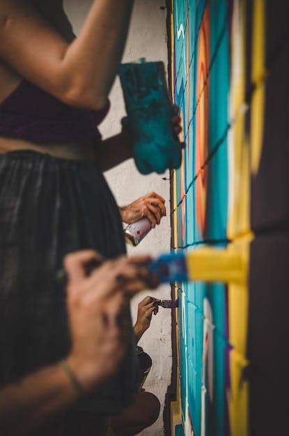 A close-up photograph of a volunteer painting intricate details on a large-scale community mural. The volunteer is holding a paintbrush and carefully adding vibrant colors to the mural, which features a diverse range of characters and symbols.